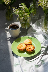 A tasty breaksfast. A plate of freshly-baked muffins with a hot cup of coffee on a white tablecloth, bouquets of lilies of the valley in glass vases on the gray background.