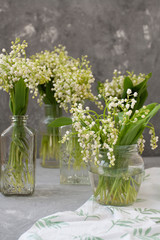 Wresh bouquets of lilies of the valley in glass vases on the white tablecloth on the gray background.