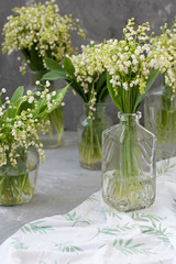 Wresh bouquets of lilies of the valley in glass vases on the white tablecloth on the gray background.