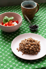 A healthy and nutritious breakfast: a rich plate of buckwheat, a cottage cheese and tomato salad with basil, a small bowl of prune jam, a hot cup of coffee on a green tablecloth background.