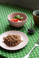 A healthy and nutritious breakfast: a rich plate of buckwheat, a cottage cheese and tomato salad with basil, a small bowl of prune jam, a hot cup of coffee on a green tablecloth background.