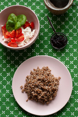 A healthy and nutritious breakfast: a rich plate of buckwheat, a cottage cheese and tomato salad with basil, a small bowl of prune jam, a hot cup of coffee on a green tablecloth background.