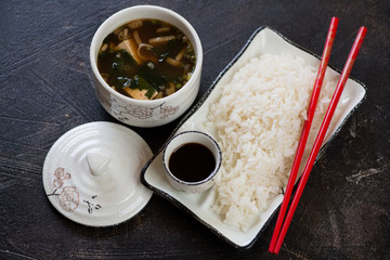 Bowl of miso soup and a plate with white rice, elevated view on a dark brown stone background, horizontal shot