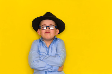 beautiful little boy in a black hat and blue shirt stands with his arms crossed isolated on a yellow background