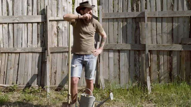 Full body portrait of bearded man farmer with shovel having fun in a farm on wooden dackground. Eco farm. Hipster farmer relaxing at nature background. Ecology concept.