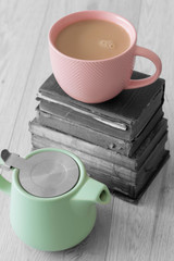 Cup of tea in a pink cup with pastel green teapot and a stack of old books with selective colour on a grey wood background