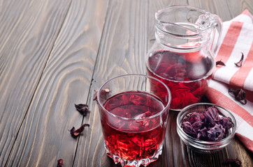 Closeup view at glass of hibiscus ice tea and jug on wooden table background