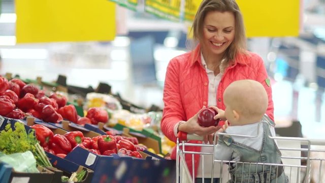 Mom and baby in the supermarket choose vegetables. A woman stretches a red sweet pepper to her one-year-old son. Healthy lifestyle, the development of babies up to a year