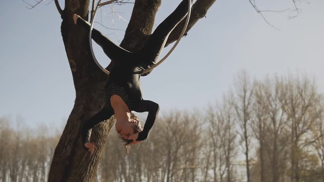 Beautiful woman doing some acrobatic elements on aerial hoop outdoors. Flexible brunette with bare feet hanging in ring for aerial acrobatics. Medium shot