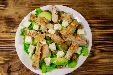 Tasty salad with fried chicken breast, green olives, feta cheese, avocado, lettuce and olive oil on wooden table. Top view