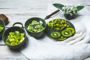on a white, wooden table top, green tableware with fresh yoghurt and pieces of kiwi fruit sprinkled with coconut flakes, next to a tiny bouquet of field flowers and leaves in green and white color