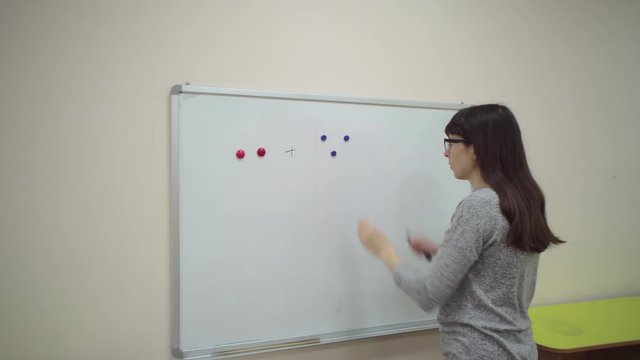 Female teacher explains rules of addition. Schoolmaster makes mathematical example on chalkboard, two red and three blue magnets through plus sign, then writes equal sign and number five.