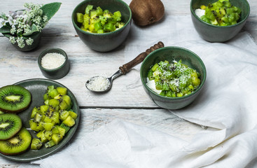on a white, wooden table top, green tableware with fresh yoghurt and pieces of kiwi fruit sprinkled with coconut flakes, next to a tiny bouquet of field flowers and leaves in green and white color