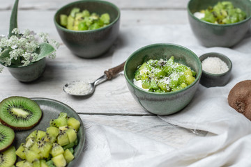 on a white, wooden table top, green tableware with fresh yoghurt and pieces of kiwi fruit sprinkled with coconut flakes, next to a tiny bouquet of field flowers and leaves in green and white color