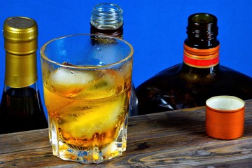 Alcoholic drink with ice on wooden table top, blue background. Strong aromatic alcoholic drink, from different types of grain - whiskey or Bourbon. Cognac or brandy - from grape must.