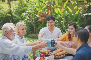 Happy big family have a lunch at outdoor in green garden. Lunch or tea time on picnic table in summer. All people have a clink glasses. Big family outdoor lunch concept.