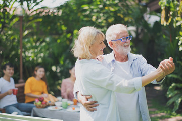Senior Caucasian couple dancing felling happy in green garden and them's family are looking in background. Grand father's birthday party in summer. Big family outdoor party concept.
