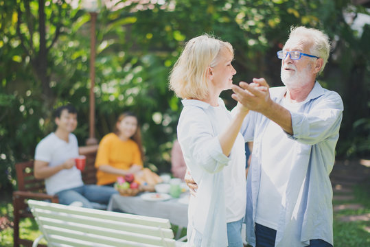 Senior Caucasian couple dancing felling happy in green garden and them's family are looking in background. Grand father's birthday party in summer. Big family outdoor party concept.
