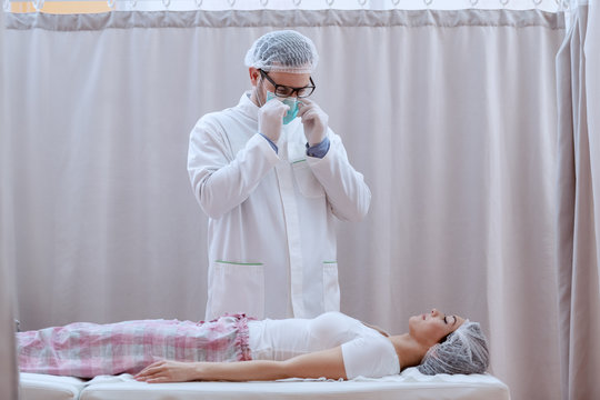 Young Caucasian Doctor In White Uniform And With Protective Mask On And Hair Net Checking On Female Patient Before Surgery.