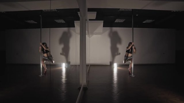 Attractive woman pole dancing at dark studio with mirror. Woman with sexy shape is dancing near the pole in slow motion. Dancer moving with pylone