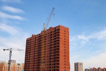 Fototapeta na wymiar Construction of a multi-storey brick residential building of red color next to which stands a crane for lifting goods and materials to the very top under a blue sky with clouds on a clear sunny day.