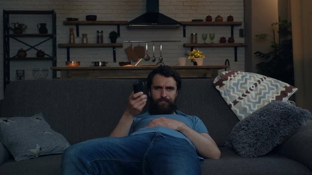 Tired man sitting on thew sofa at home after work. Holding remote control tv and switching channels. Looking for TV program.