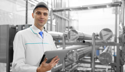 Portrait of man in a white robe and a cap standing in production department of dairy factory with grey tablet