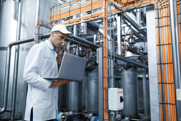 Portrait of man in a white robe and a cap standing in production department of dairy factory with laptop