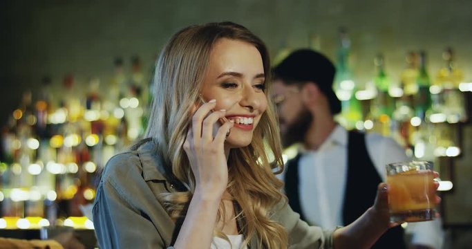 Close up of the happy Caucasian blond pretty girl drinking cider and having a nice talk on the mobile telephone at the bar.