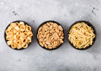 Ready salted potato rings with salt and vinegar sticks and roasted peanuts as classic snack in black bowls on light background.