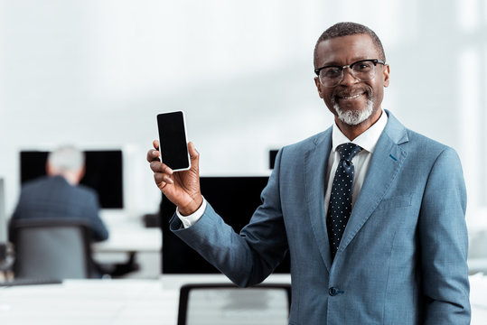 selective focus of happy african american businessman holding smartphone with blank screen in office