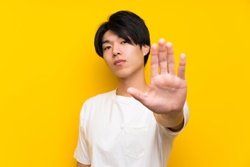 Asian man over isolated yellow wall making stop gesture