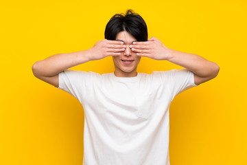 Asian man over isolated yellow wall covering eyes by hands