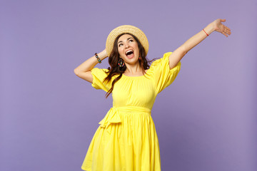 Portrait of funny screaming young woman in yellow dress, summer hat rising hand, looking up isolated on pastel violet wall background. People sincere emotions, lifestyle concept. Mock up copy space.