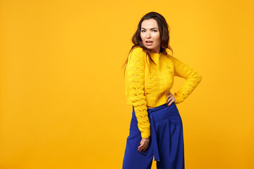 Portrait of passionate young woman in sweater, blue trousers standing with arm akimbo on waist isolated on yellow orange wall background. People sincere emotions lifestyle concept. Mock up copy space.