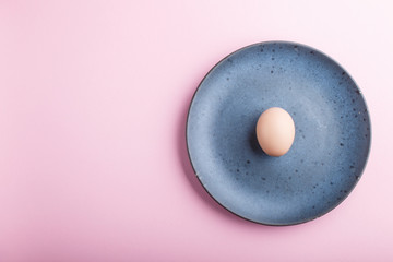 Raw uncooked single egg on a blue ceramic plate on a pink pastel background. top view.