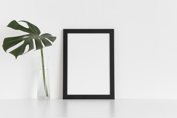 Black frame mockup with a monstera leaf in a glass vase on a white table. Portrait orientation.