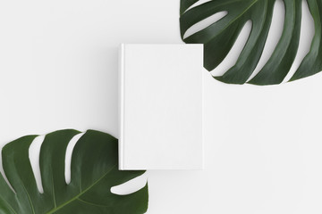 Top view of a white book mockup with monstera leaf decoration on a white table.