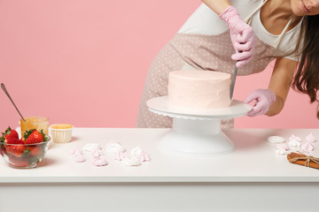 Close up cropped chef cook confectioner or baker in white t-shirt cooking at table isolated on pink pastel background in studio. Cream application cake making process. Mock up copy space food concept.