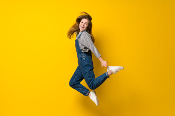 Young girl with overalls jumping over isolated yellow background
