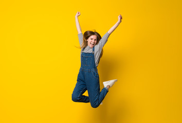 Young girl with overalls jumping over isolated yellow background