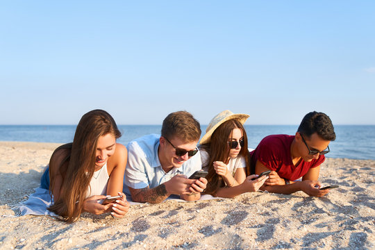 Group of millenials using smartphones laying together on beach towel near sea on summer sunset. Young people addicted by mobile smart phones. Always connected generation communicate via internet.