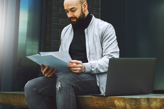 Young bearded business man working on laptop while sitting on bench outdoors. Man holds paper documents in his hands. Lifestyle.
