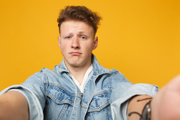 Close up selfie shot of displeased young man in denim casual clothes looking camera isolated on yellow orange wall background in studio. People sincere emotions lifestyle concept. Mock up copy space.