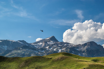 Obraz premium Over a green alpine pasture in a blue sky with white clouds a small black helicopter is flying, French Alps, National Park Vanoise. 