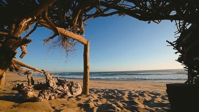 Relaxing slow motion view of idyllic, paradise island sandy beach & slow moving ocean waves from inside handmade log & tropical leaves beach cabin shelter.