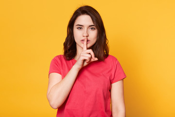 Closeup portrait of secretive young woman keeps finger on lips asking shh, quiet, silence looking directly at camera isolated over yellow background. Human expressions, emotion, body language.