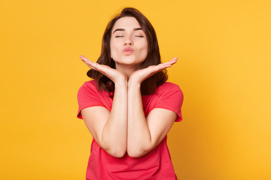 Close up portrait of pretty Caucasian woman makes kiss with lips, keeps both hands under chin, wearing casual red t shirt, poses with closed eyes over yellow wall. Happyness and gladness concept.
