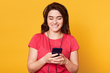 Portrait of beautiful dark haired listening to music on mobile phone, standing against yellow background wearing red t shirt, enjoying to spend free time. Blank space for promotion or advertisment.