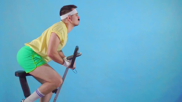 Funny young man from the 80's with a mustache and glasses on exercise bike on a blue background